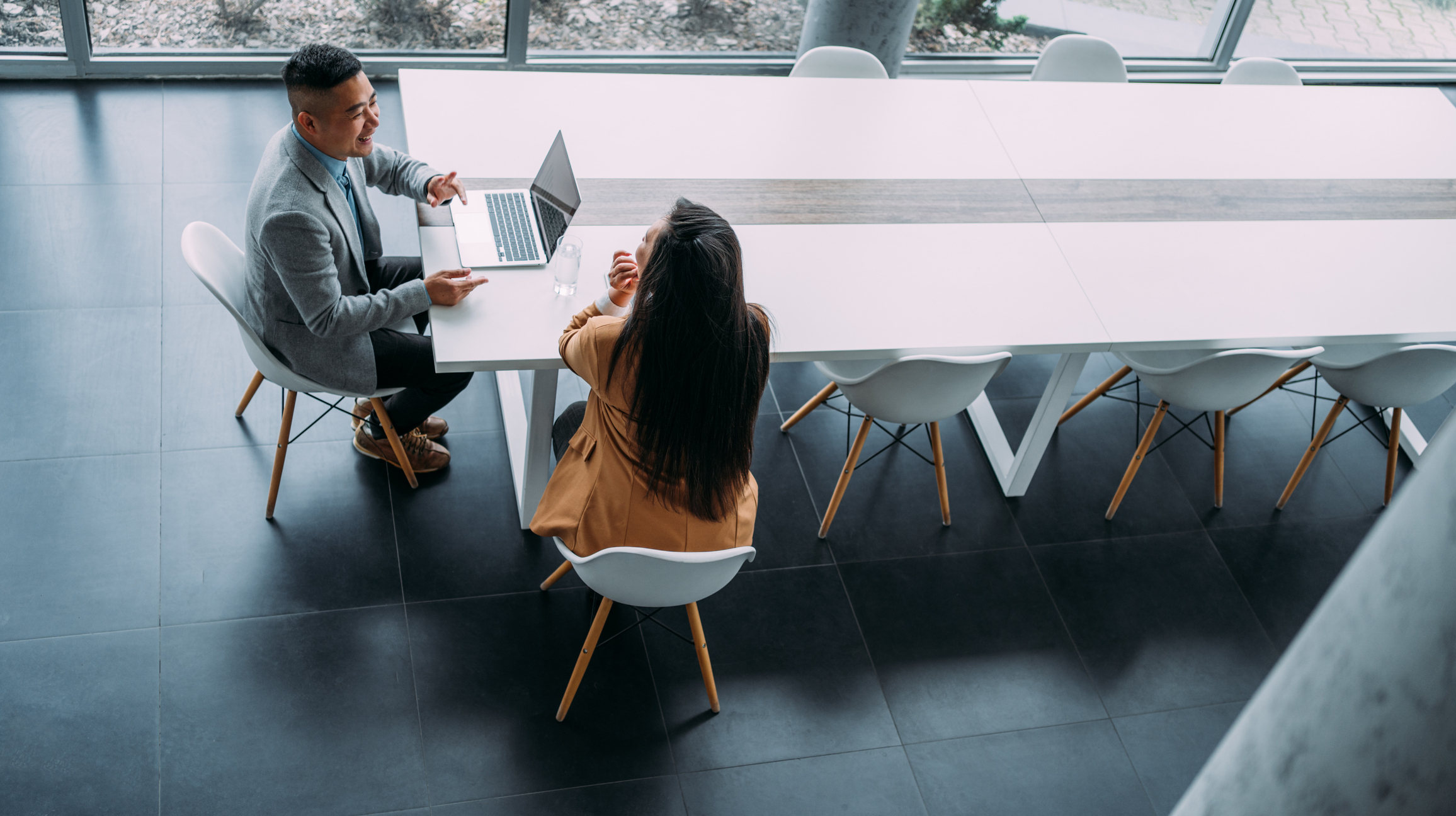 Above view of a two confident business persons sitting on a conference table in the office and sharing ideas. Businessman and businesswoman in meeting using laptop and discussing business strategy. Business coworkers working together in the office.